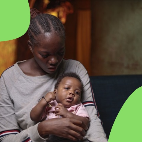 A young woman with braided hair holds an alert baby in her lap indoors. She looks down calmly, with green shapes framing the softly lit background.